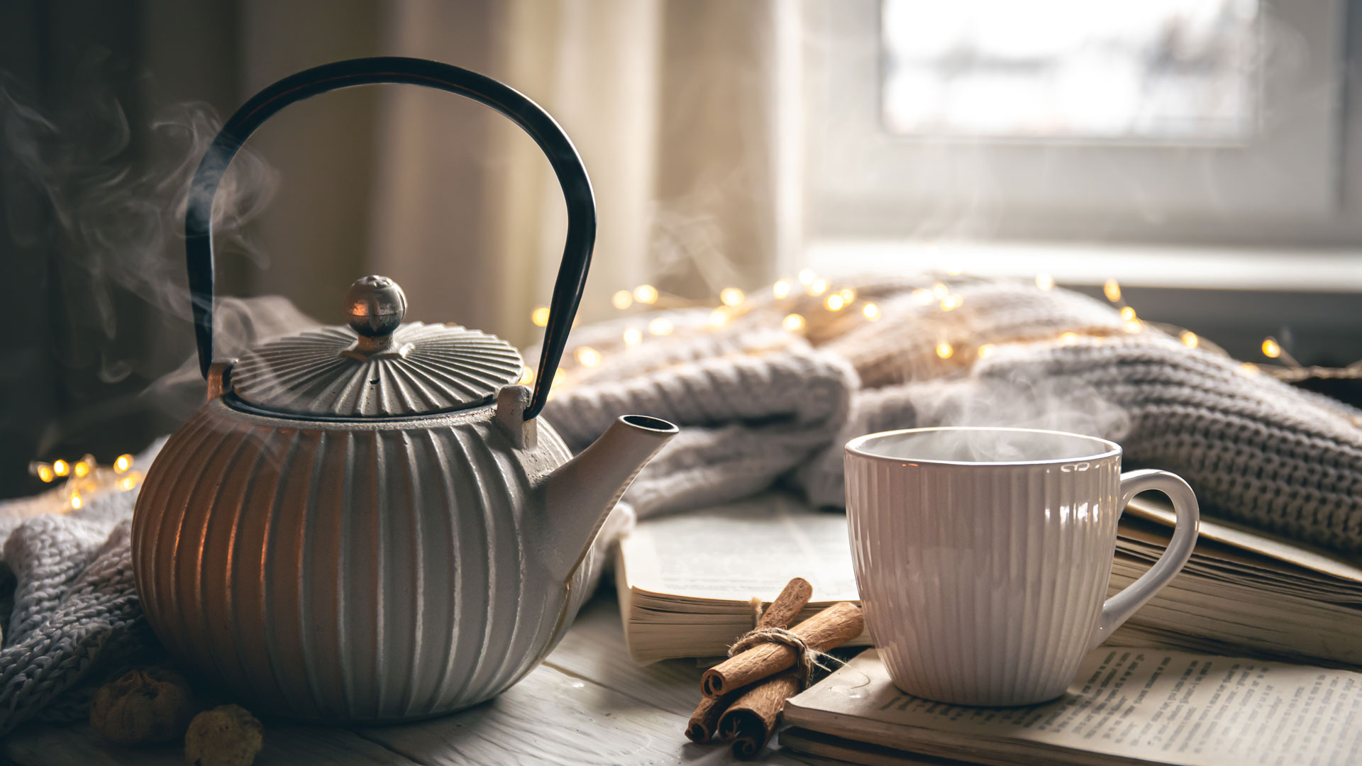 Gemütliche Winteratmosphäre: Dampfende Teekanne mit Teetasse auf einem Buch mit einer Wolldecke und einer Lichterkette im Hintergrund, durch ein Fenster kommt warmes Winterlicht.