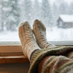 Im Vordergrund sieht man Füße in dicken Wollsocken auf der Fensterbank mit Blickrichtung aus dem Fenster. Draußen fallen Schneeflocken auf eine winterliche Landschaft mit Bäumen und einem kleinen Haus. Drinnen sorgen eine Tasse mit dampfendem Getränk, eine Lampe und eine Strickdecke für eine ruhige, warme Atmosphäre.