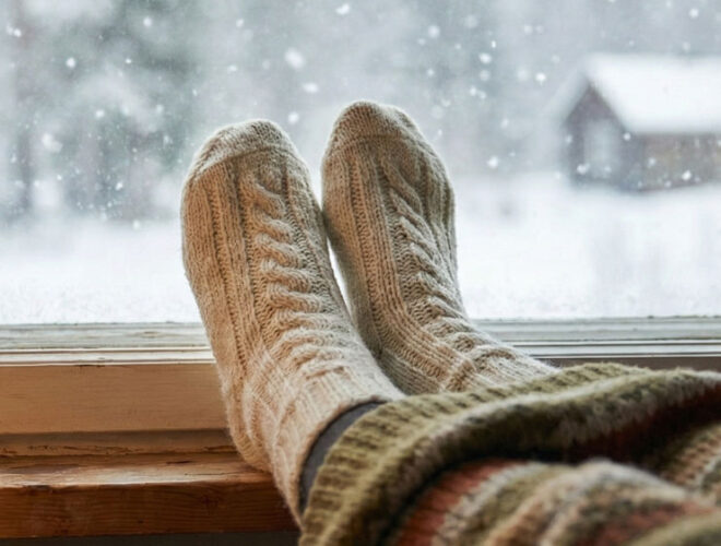 Im Vordergrund sieht man Füße in dicken Wollsocken auf der Fensterbank mit Blickrichtung aus dem Fenster. Draußen fallen Schneeflocken auf eine winterliche Landschaft mit Bäumen und einem kleinen Haus. Drinnen sorgen eine Tasse mit dampfendem Getränk, eine Lampe und eine Strickdecke für eine ruhige, warme Atmosphäre.